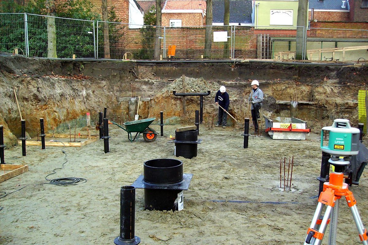 Uitgegraven bouwput met twee medewerkers met veiligheidshelm, kunststof inspectieputten, stempels en een rotatielaser op statief.
