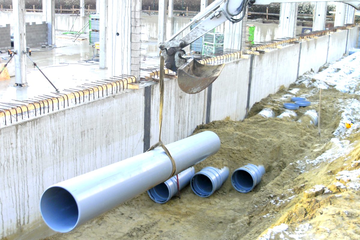 Graafmachine van Schockaert die een grote grijze rioleringsbuis in de sleuf laat zakken.