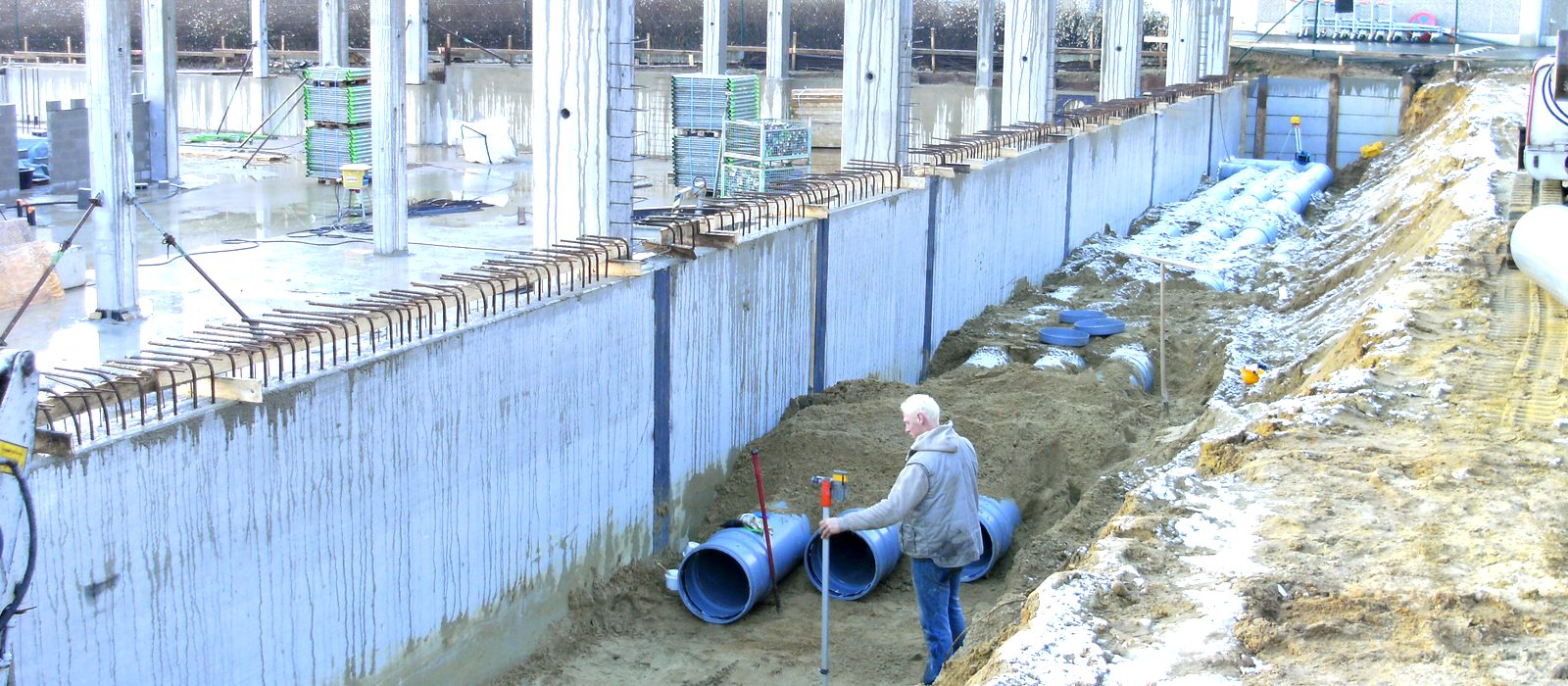 Werf van Schockaert met een sleuf naast een betonnen kelderwand, blauwe rioleringsbuizen van grote diameter en een medewerker met meetlat.