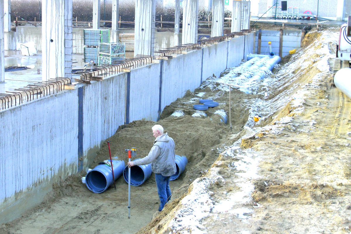 Sleuf naast een betonnen kelderwand met blauwe rioleringsbuizen van grote diameter, klaar om te leggen.